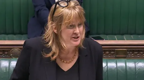United Kingdom Parliament A woman with long blonde hair stands in the House of Commons dispatch box with green chairs behind her. She is wearing a black suit, shirt and gold necklace.