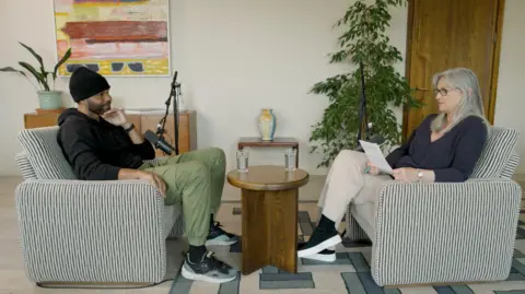 Made by Dyslexia A man on the left and a woman on the right are sitting in chairs facing each other. They both wear dark tops. The woman has gray hair and wears glasses.