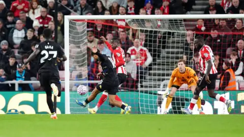 PA Media Elimane Ndiaye wears a black kit. He can be seen kicking the ball into the goal on a football field while a player wearing red and white shirts tries to intercept it. They are on a green soccer field and the goalkeeper is wearing an orange jersey. 