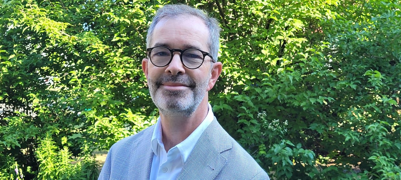 A man stands in front of greenery smiling in glasses