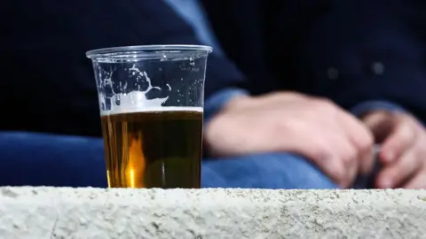 Getty Images A pint of beer in a plastic cup on the wall. A person can be seen sitting in the background.