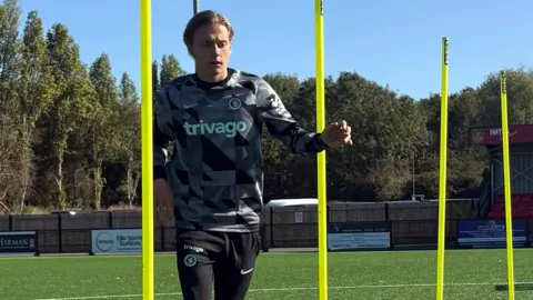 George Edwards wears a gray and black plaid shirt and black Nike sweatpants. He