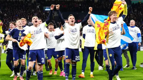 PA Media Football players celebrate on the pitch, wearing white shirts over their team kits and carrying Scottish flags.