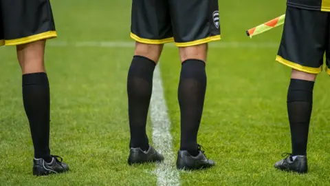 Getty Images The lower bodies of three football match officials standing on the grass. The photo was taken from behind as they wore a black and yellow ensemble.