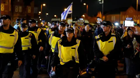 Palestinian Authority Media Several police officers wearing hats and hoods stand in front of protesters carrying Israeli signs and flags on a city street at night. Read some signs "Keep anti-Semitism out of football". 