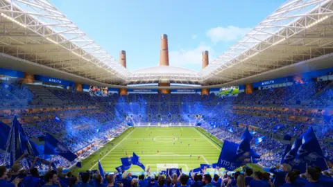 The city of Birmingham inside the ground has a sea of ​​blue as fans sit in this artistic impression. The field can be seen with the players on it. The advertising screen surrounds the upper ring around the floor, which is partially covered by a white ceiling.