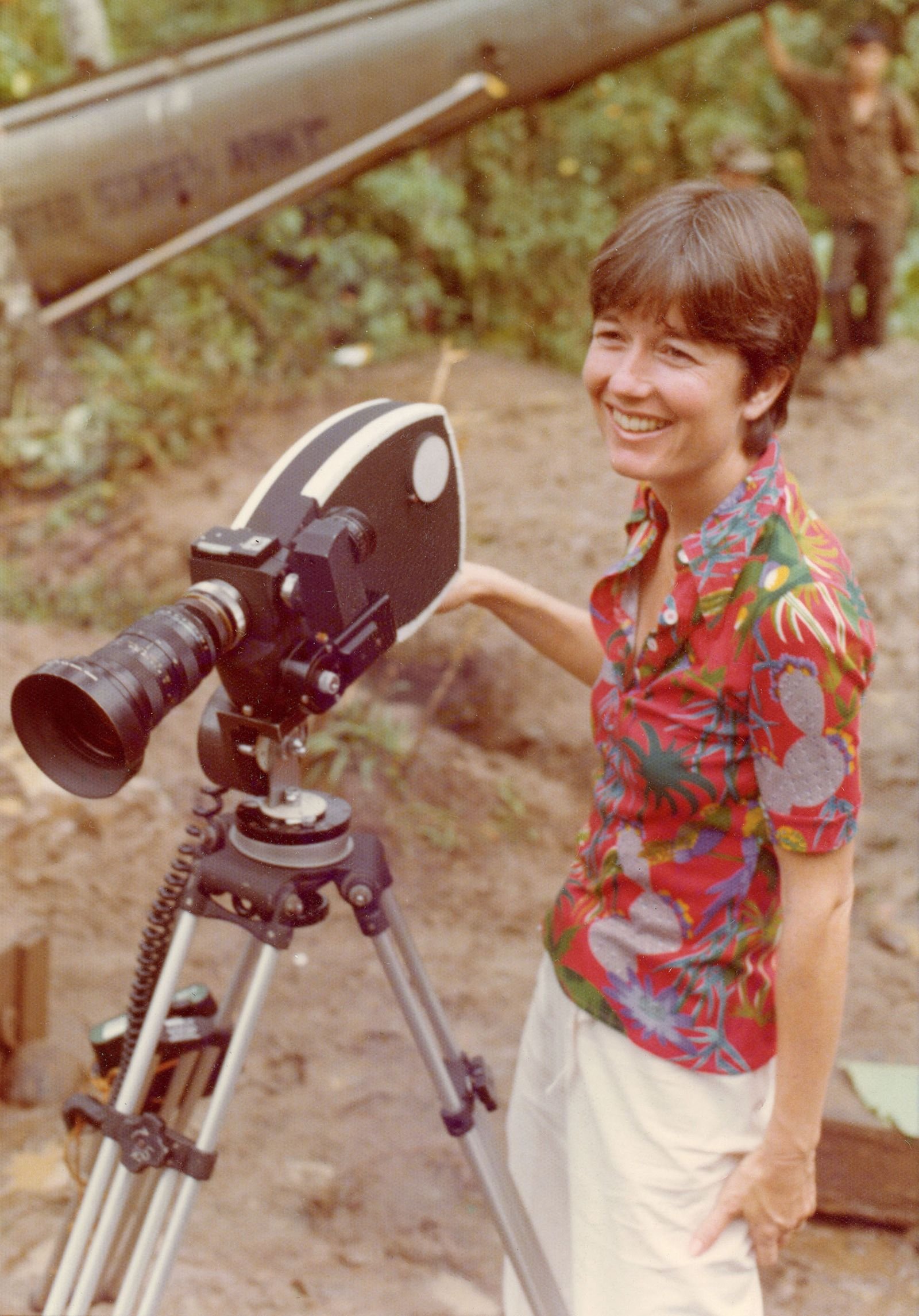 A character wearing a short-sleeved button-up stands next to a camera on a tripod and smiles from a distance.