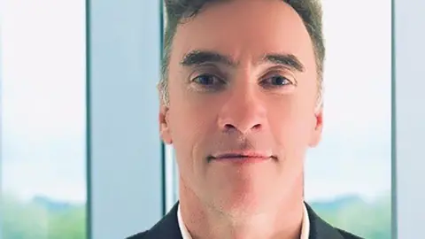 Unidentified Paul Doyle, with gray hair styled in a bob, smiles at the camera while wearing a black suit jacket and white open-collar shirt.