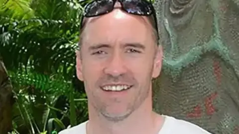 Paul Doyle smiles while holding black sunglasses on his head. He is wearing a white shirt.