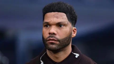Alex Livesey - Danehouse/Getty Images A man with dark hair and a beard looks away from the camera. The background is blurred in light blue.