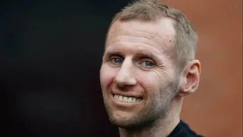 PA Media Rob Burrow smiles for the camera with short brown hair, brown beard and black top. The background is also black and brown.