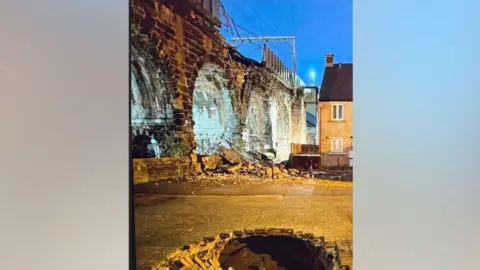 Network Rail A photo showing damage to a bridge. A section of the barriers that run along the top of the bridge appears to have collapsed and a pile of rubble can be seen underneath. A large hole can be seen in front of the bridge