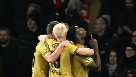 Reuters Crystal Palace player Mark Guehi looks to the sky and points. Many of his teammates hug him from behind. They all wear gold shirts. 