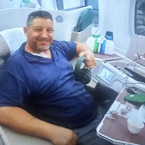 Erez Hadari Erez Hadari is shown sitting in a plane - in what looks like first or business class - with a blue top and grey trousers, and is smiling, holding headphones