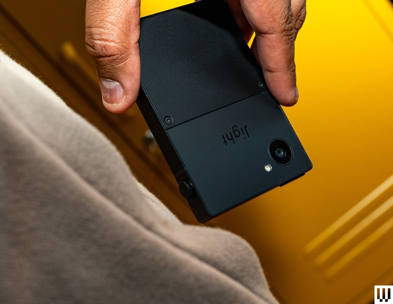 A hand holds the Light Phone III, a small, black, square device pulled from a side pants pocket