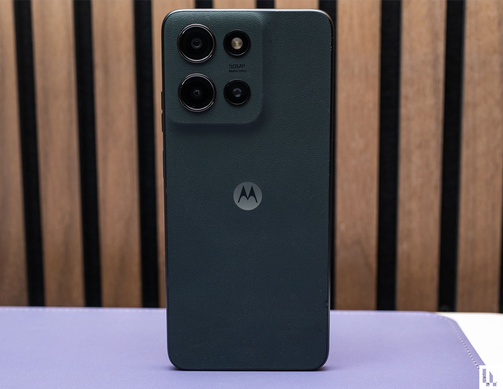 Moto G 2025, a black mobile phone with a back showing four cameras placed on a purple mat with a wooden wall in...