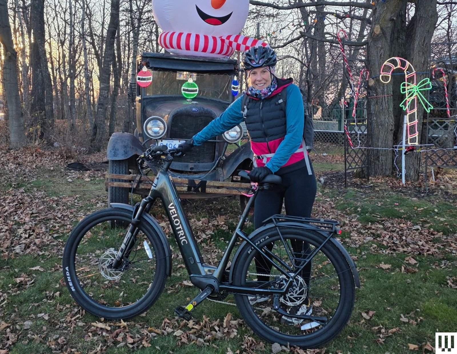 A person stands next to a black Velotric Discover 2 electric bike with holiday decorations in the woods behind