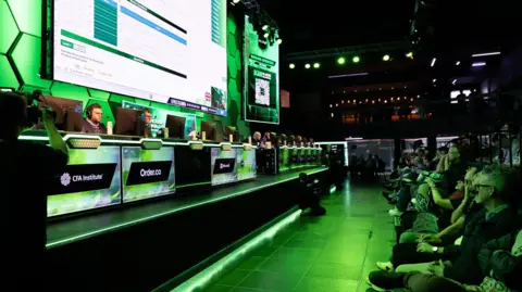 MEWC An arena featuring a stage raised to the left, and the crowd to the right.
Those on the left sit at computer desks with large monitors, focused on the Excel challenges, with a stream of it live on a massive screen behind them. The crowd on the right watch on enthusiastically, covered in the green lighting cast by the arena.