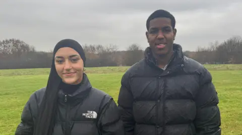 Zainab wears a black hijab and a black coat from The North Face. Jayden is wearing a black puffer coat. They stand side by side in a field with trees in the distance. 
