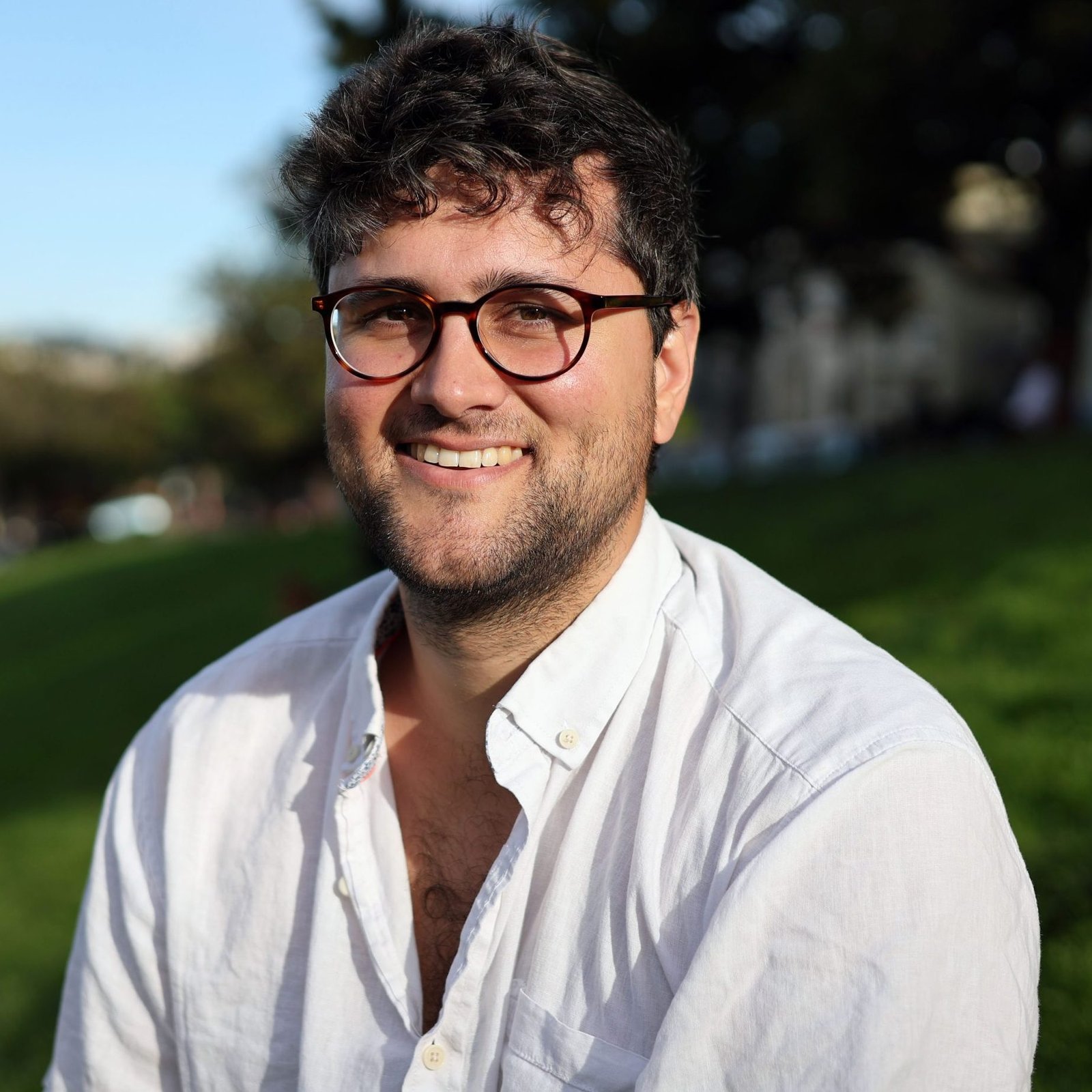 A man with glasses and a beard smiles while sitting on grass in a park, wearing a white shirt.