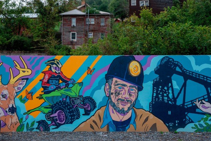 A mural features a deer, an ATV rider, a miner with a headlamp, butterflies, and a coal mining structure against a colorful background.