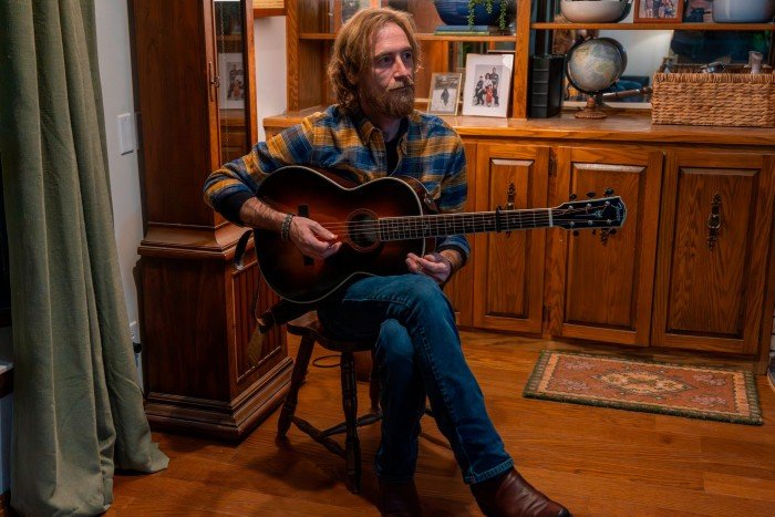 Curtis Lovejoy sits in a wood-paneled room playing an acoustic guitar, looking thoughtfully to the side.