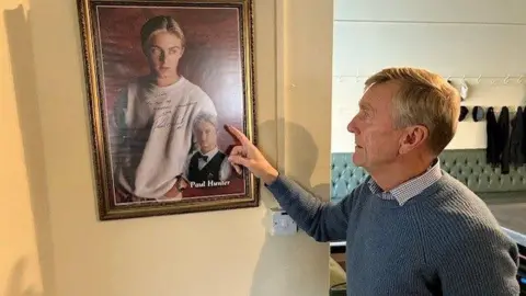 David Spereall/BBC A man in a blue jacket points to a signed photo of Paul Hunter on the wall.