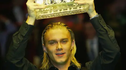 Getty Images Snooker player Paul Hunter holds the cup high out of the shot. His blond hair is braided and he wears a black shirt.
