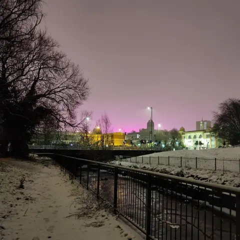 EliP/BBC Weather Watchers There is a river in the foreground and illuminated buildings can be seen in the background. The sky has a strange tint of pink and purple.
