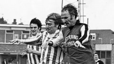 Kettering Town FC Black and white photo of Derek Duggan wearing the famous Kettering Town FC kit. I have "Kettering tires" Printed on the front. Dogan struggles for the ball with two opposition players.