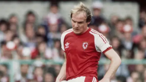 Getty Images Terry Yorath on the Wales set in 1980. He looks at a soccer ball as he kicks it. The crowds at the stadium can be seen blurred in the background.