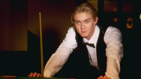 Getty Images A young Paul Hunter poses for a photo while leaning against a snooker table. Cue stands next to his right hand. He has floppy blonde hair and wears a tie, white shirt, and jacket.