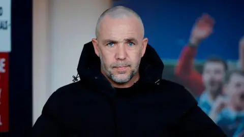 SNS Dougie Imrie - Man with shaved head, gray beard, mustache and black jacket, on the touchline during a football match