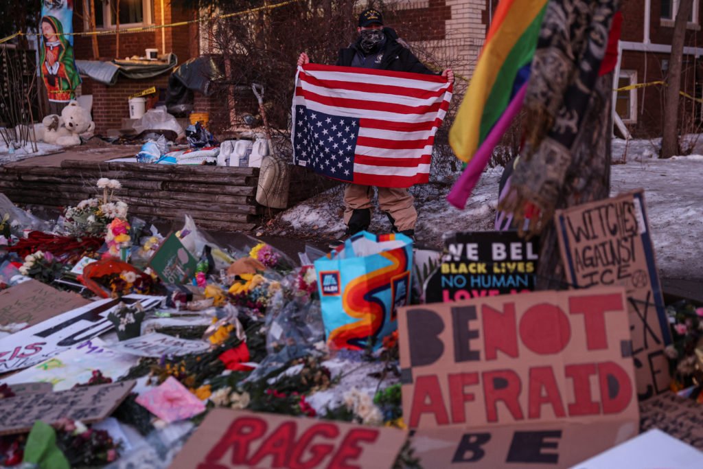 A person holds an upside-down American flag while standing next to a makeshift memorial at the scene of Renee
