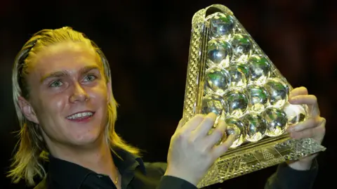 Getty Images Paul Hunter lifts the Masters Cup. The cup is a glass sculpture of a triangle of red balls. Hunter has braided blonde hair.