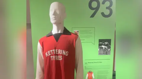 Brian Lewin is a model on display wearing a 1976 Kettering Town football kit. It is red with a black collar and white sleeves. "Kettering tires" Printed with white line down the middle. Behind the mannequin is a green screen showing the history of the shirt.