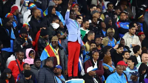Reuters Michel Ngcuka Mboladinga dressed as Patrice Lumumba, wearing a blue jacket and red shorts, his hand raised among Congolese fans in a stadium in Rabat.