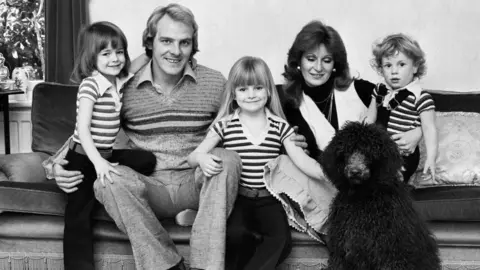 Getty Images Black and white photo of a family with three children, a father and a mother. Everyone sat on the sofa with a fluffy black dog.