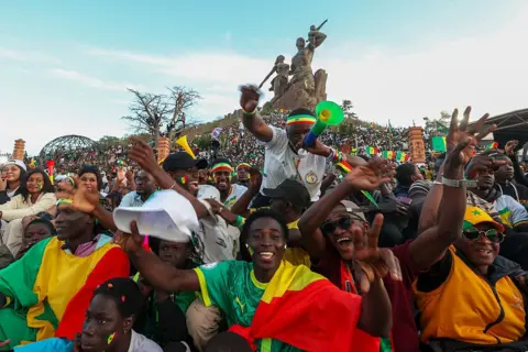 Andalusia via Getty Images Senegalese fans wearing clothing and accessories in the colors of the national flag gather around the African Renaissance Monument, chanting and singing in support of their team.