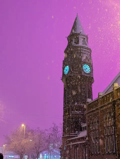 GLMCC Pink sky above the clock tower in Birmingham city centre. The clock face is illuminated and heavy snow can be seen falling around it. 
