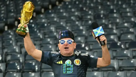 Getty Images A man dressed in Scotland jersey holds a replica of the gold World Cup trophy above his head. He wears a tartan hat, a Scotland ribbon, and sunglasses with salters on the lenses. He