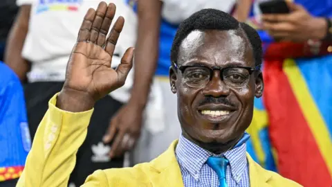 AFP/Getty Images Michel Ngcuka Mboladinga dressed as Patrice Lumumba, wearing a yellow jacket, blue shirt and tie, raising his hand as if waving in the stands at the Africa Cup of Nations.