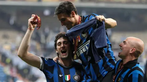 Reuters Inter Milan players Diego Milito, Javier Zanetti and Esteban Cambiasso celebrate after winning the Champions League in 2010. Their kit has blue and black stripes and is labeled with Pirelli in white. Zanetti is carried aloft by the other players.