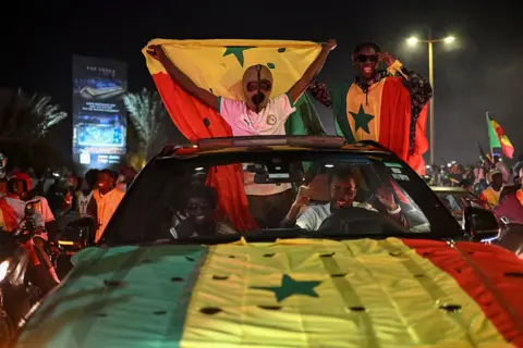 AFP via Getty Images Senegalese football fans celebrate at the African Memorial Square in Dakar. A fan wearing a Mexican wrestling mask waving a Senegalese flag stands across the roof of a car that also has the flag on its hood.