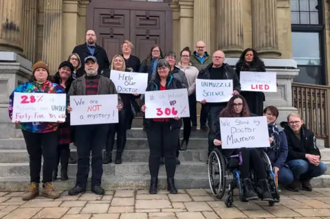 Global News Jillian Lucas and other patients stand in front of a government building holding signs saying 