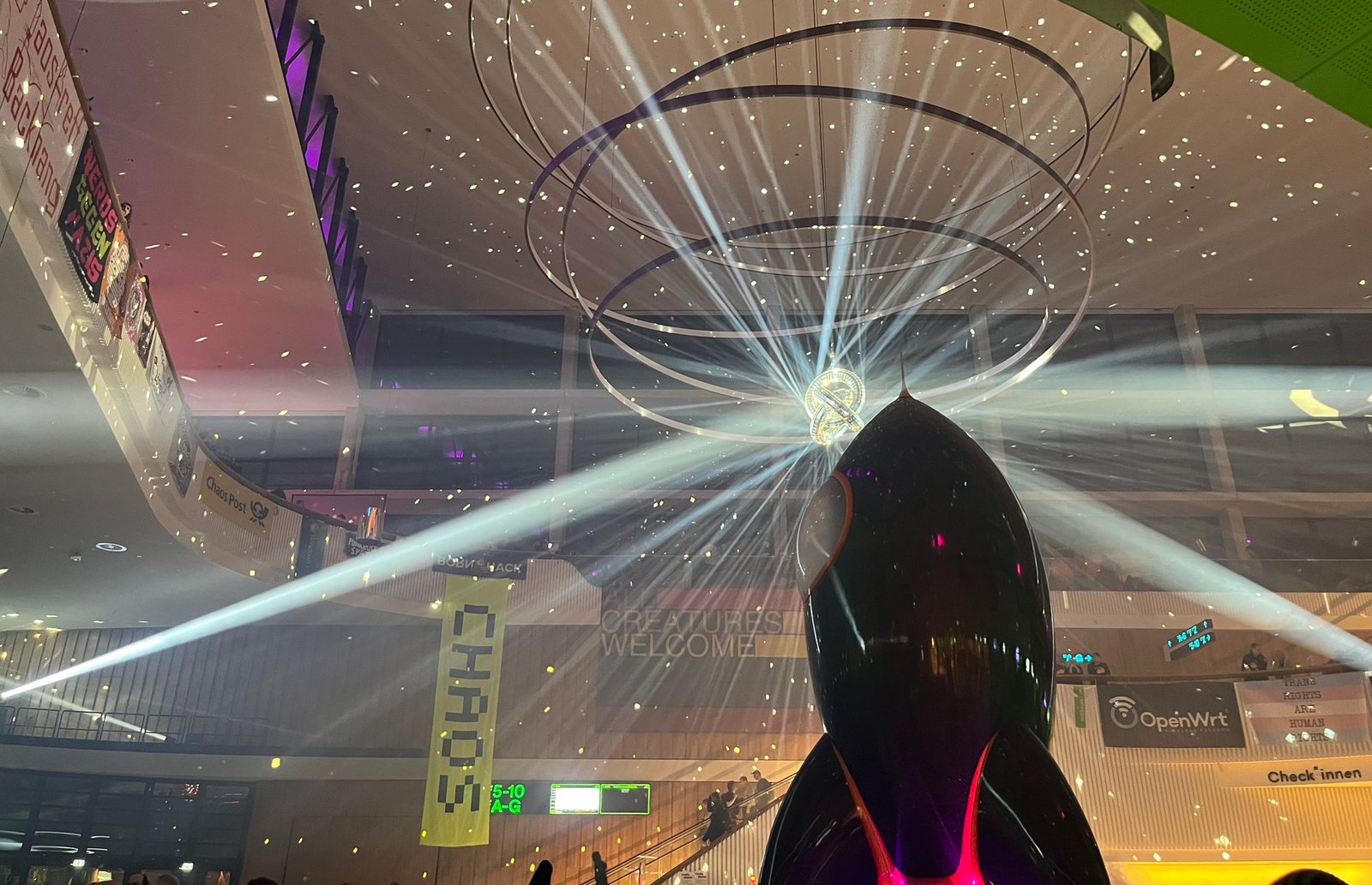 The "Fairy Dust" rocket installation at the Chaos Communication Congress (39C3), showing a large black rocket sculpture with disco "rings" behind its tip emitting dramatic light beams in all directions. The main hall of the convention center surrounds it with banners including "CHAOS", "ALL CREATURES WELCOME", "OpenWrt", and "TRANS RIGHTS ARE HUMAN RIGHTS".