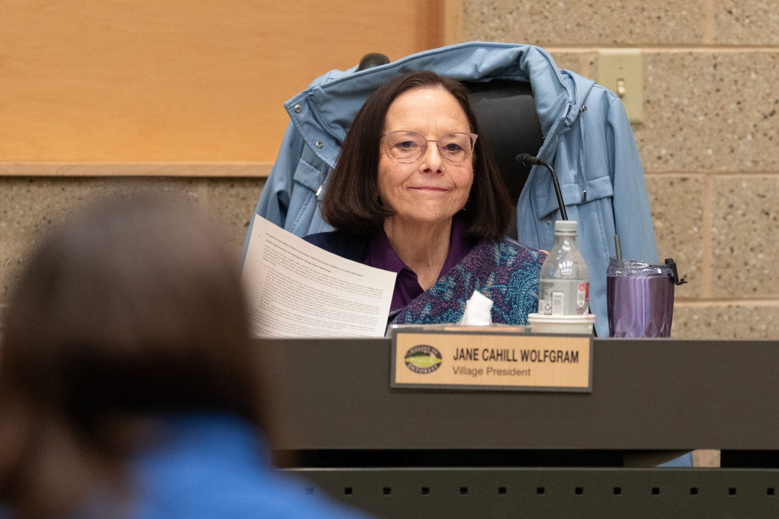 A woman sits at a desk with papers, a water bottle, and a purple cup. A nameplate reads Jane Cahill Wolfgram, Village President. A coat hangs on the back of her chair.