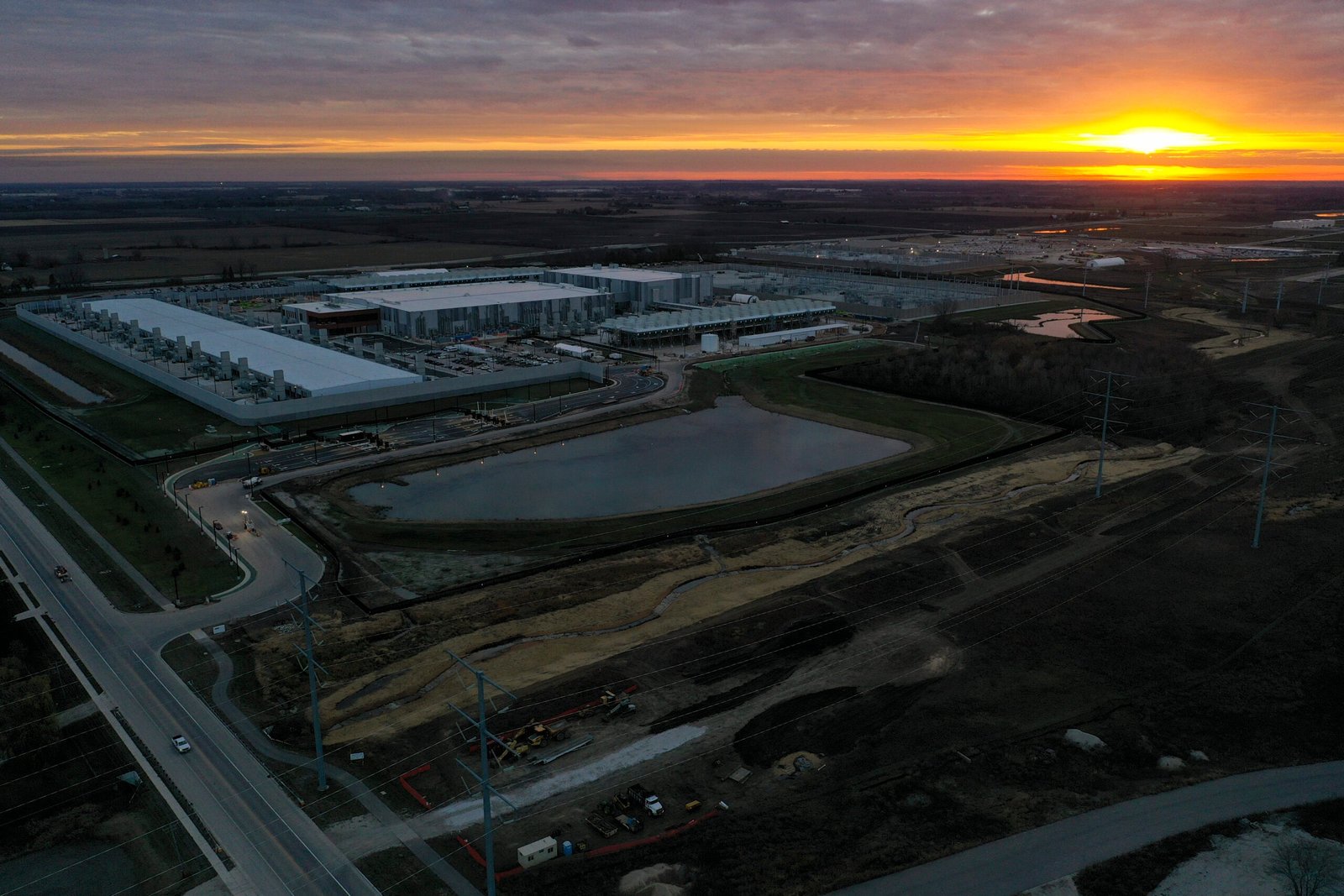Aerial view of an industrial facility with large buildings, parking lots, a pond, roads, and surrounding fields at sunset.