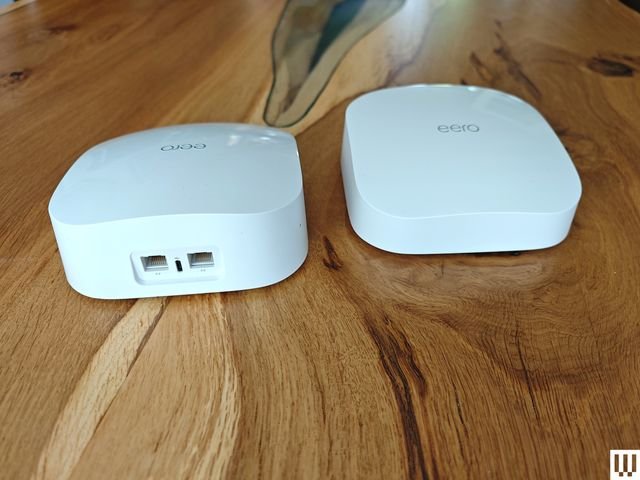 Eero 6 Mesh System, two white rectangular devices on a wooden table, one facing forward and one facing back to show the ports