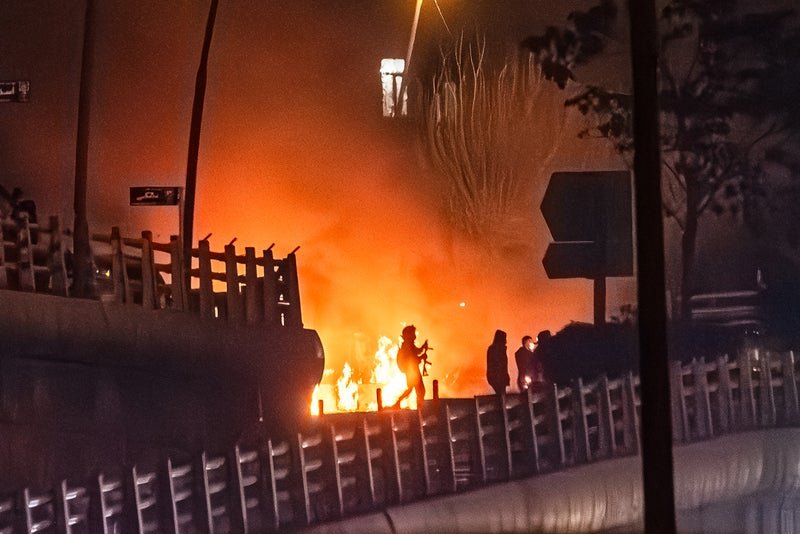 Silhouettes of people near a fire on a bridge, with smoke and orange flames illuminating the scene at night.